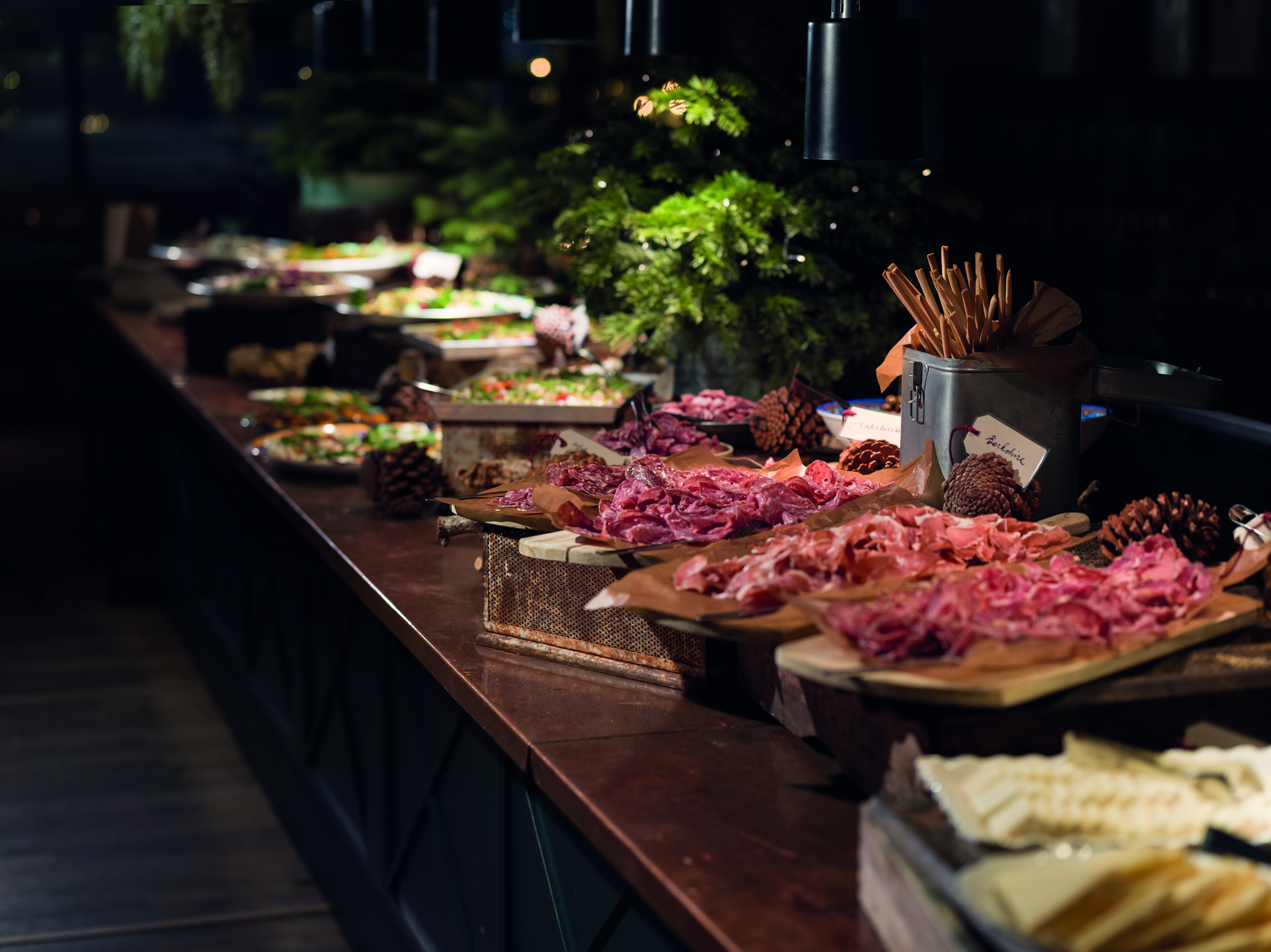 A Christmas buffet with various dishes, including cold cuts and bread, neatly arranged on a dark wooden table decorated with pine branches and cones