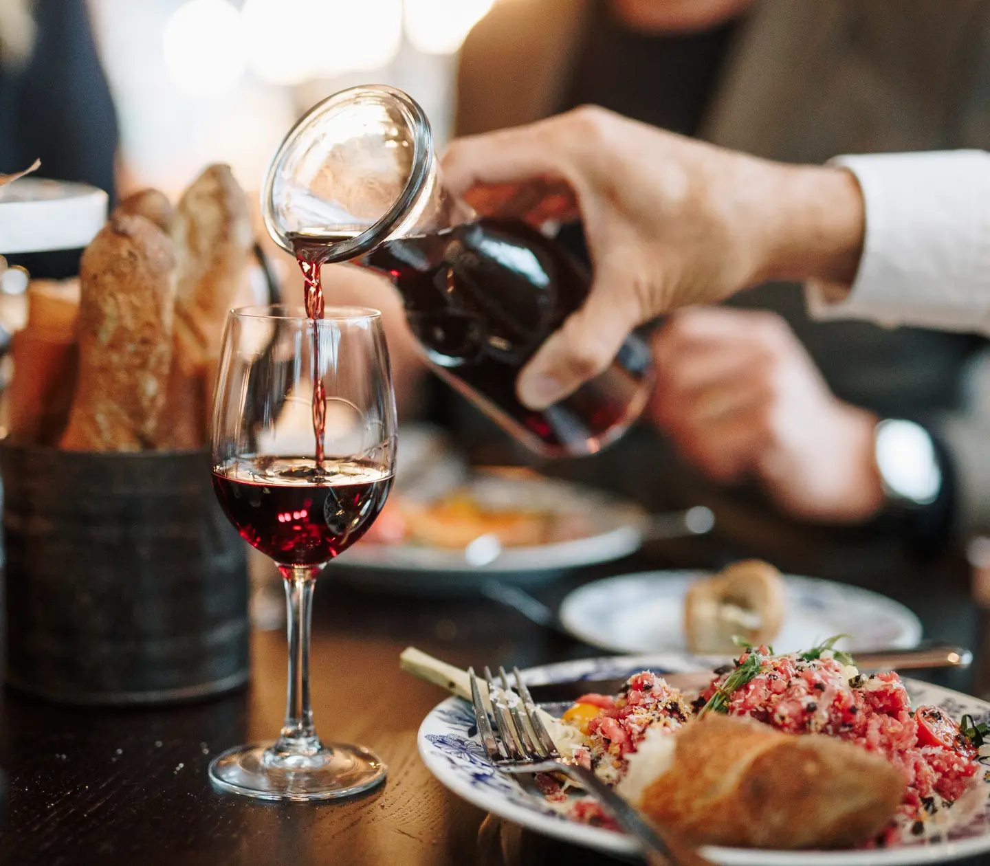 Person häller rött vin i vinglas vid ett dukat bord med maträtter och nybakat bröd på en restaurang